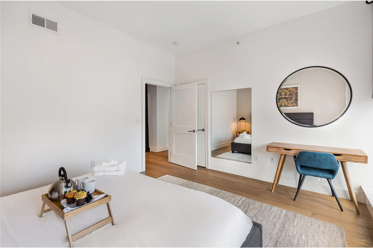 A serene bedroom with a bed dressed in crisp white linens, a breakfast tray, a round mirror, and a desk with a blue chair placed near the window. The scene evokes a sense of relaxation and comfort, with the added bonus of a dedicated workspace for guests who need to work remotely.