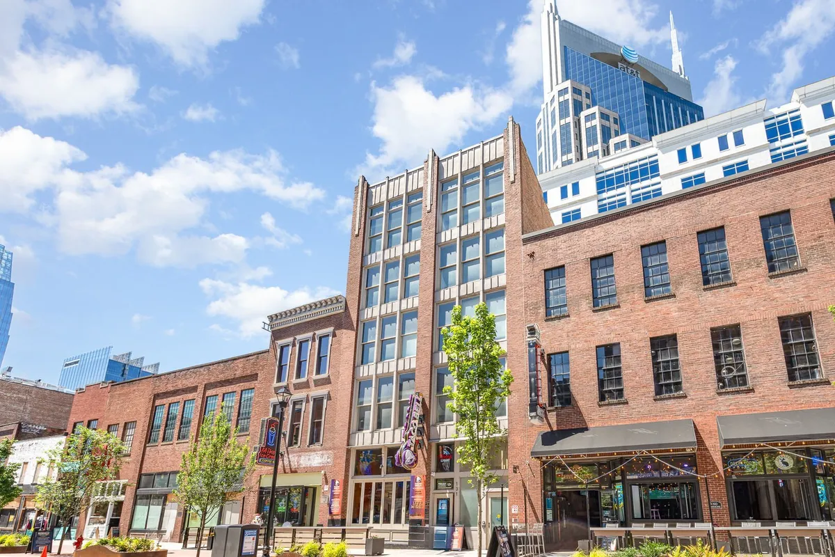 The image showcases the historic Saddlery building's charming façade set against the backdrop of modern Nashville skyscrapers. The bustling streetscape is lined with trees and features an inviting outdoor dining area, capturing the energy and vibrancy of downtown. The scene is alive with locals and visitors enjoying dining, shopping, and strolling through the lively neighborhood.