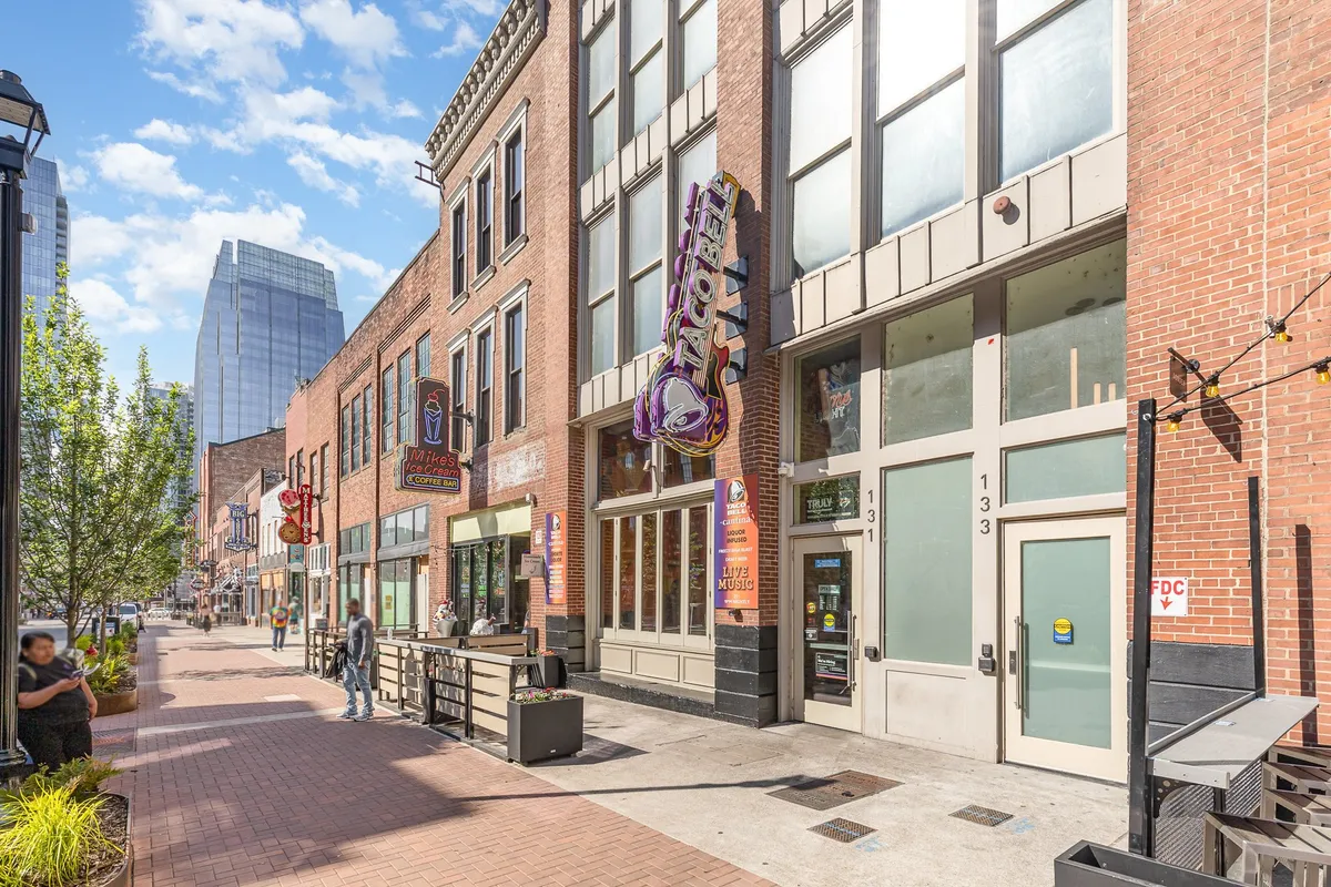 This exterior shot shows the vibrant downtown streetscape with historic brick buildings, sidewalk cafes, and the recognizable Taco Bell signage, capturing the lively urban spirit that surrounds the Saddlery building. The scene is energetic, with people dining, shopping, and enjoying the neighborhood's unique character.