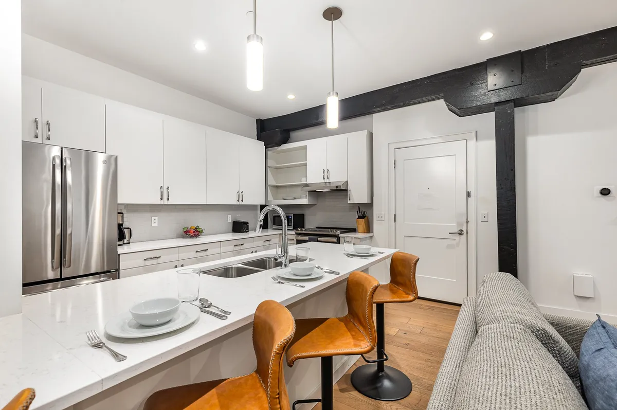 A bright, modern kitchen interior with a wide kitchen island, stainless steel appliances, open shelving, and bar stools. The setup is perfect for casual dining, with enough space for several people to gather, cook, and celebrate together.