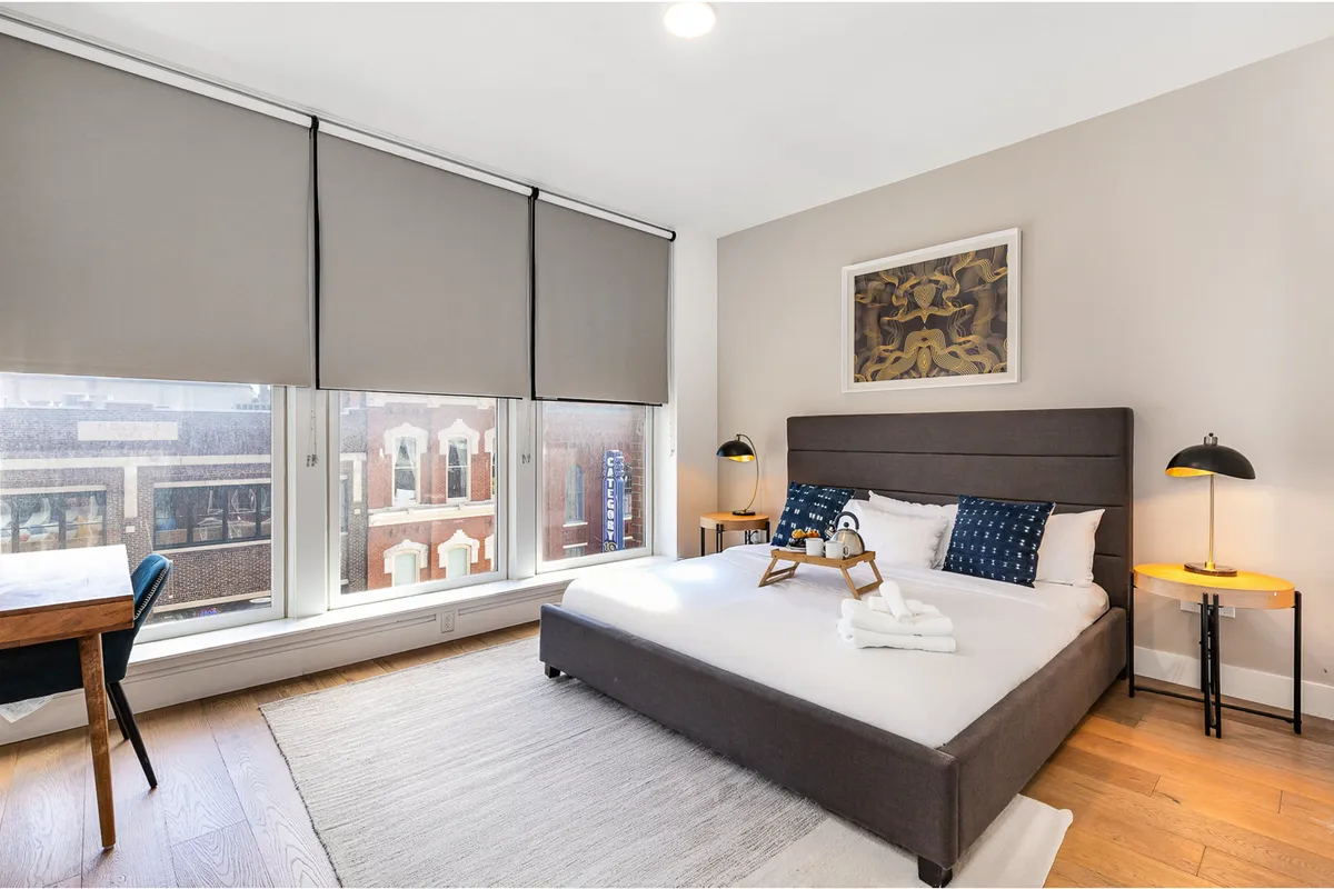 A cozy bedroom featuring a bed with decorative pillows, stylish artwork on the wall, a wooden table and chair serving as a workstation, and large windows with shades letting in natural light. The overall mood is serene and inviting, ideal for both relaxation and productivity.