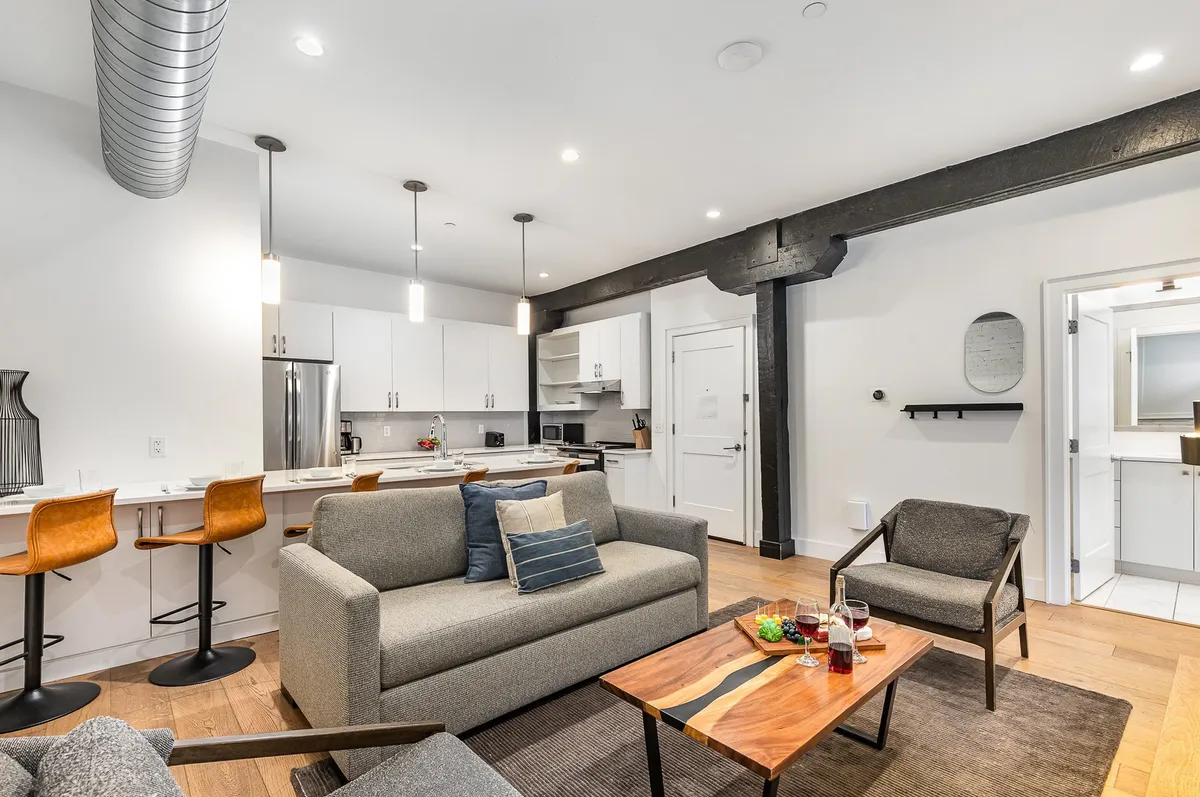 A modern, cozy living room with a plush sofa, coffee table, and adjacent kitchen area with bar stools. The setup is perfect for intimate dinner parties, casual brunches, or a quiet movie night.