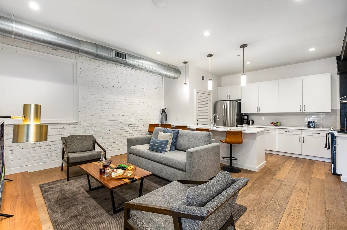 A cozy, contemporary living area with a sofa, coffee table, kitchen island with bar stools, and stylish lighting. The open layout merges socializing, relaxing, and cooking, making it ideal for gatherings or quiet moments alike.