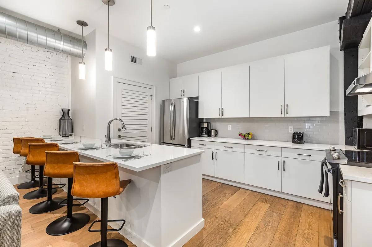 A welcoming, contemporary kitchen with a large island and bar stools, stainless steel appliances, white cabinetry, and an exposed brick wall. The space is clearly set up for entertaining and socializing while preparing meals, reflecting a blend of style and functionality.